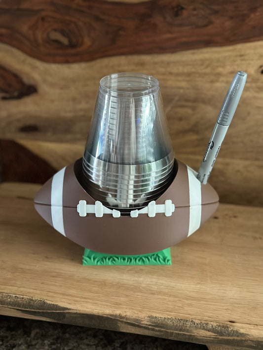 Football-shaped cup holder with a clear cup and pen on a wooden surface