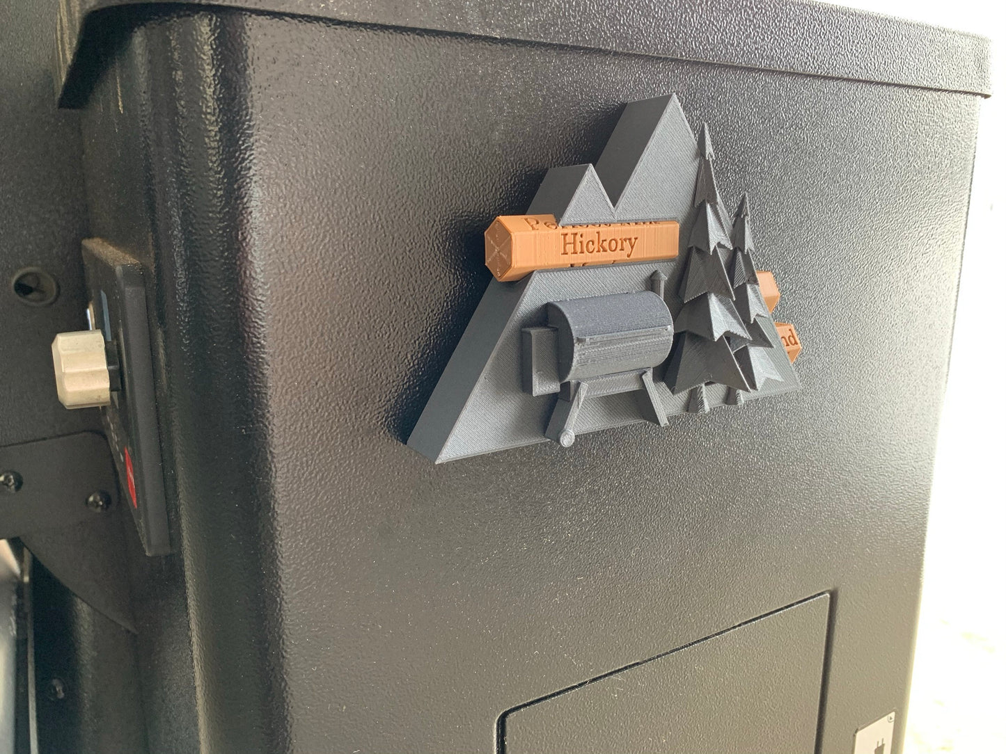 Pellet label magnet installed on a smoker hopper, showcasing its unique mountain scene design with trees and a grill and secure magnetic functionality.