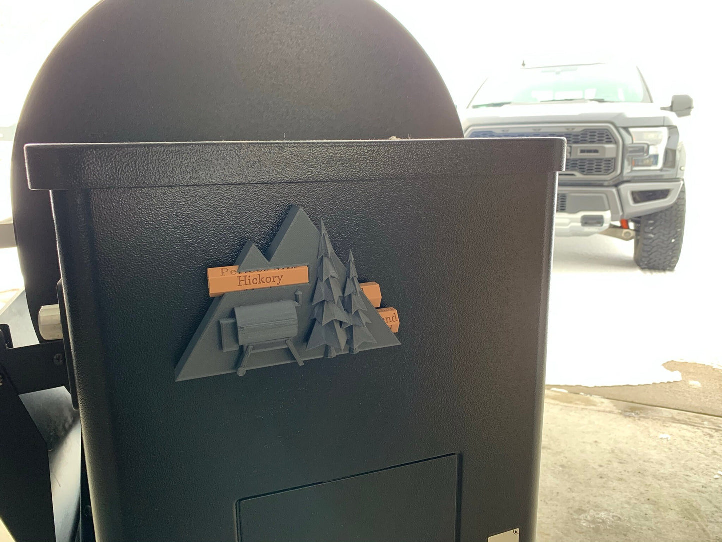 Pellet label magnet installed on a smoker hopper, showcasing its unique mountain scene design with trees and a grill and secure magnetic functionality.